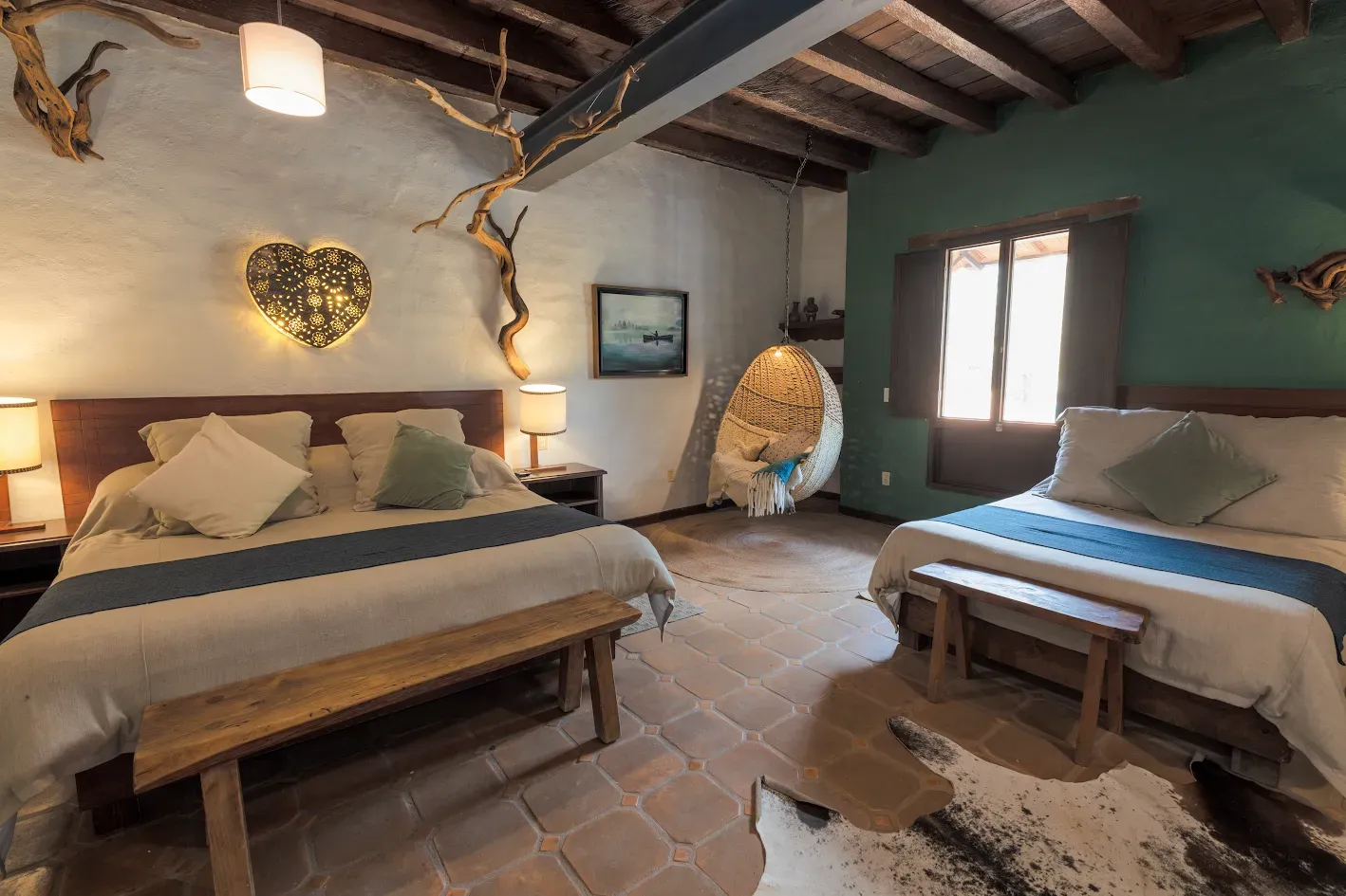 Habitación rústica con dos camas, vigas de madera, iluminación cálida y una gran ventana en paredes de piedra.