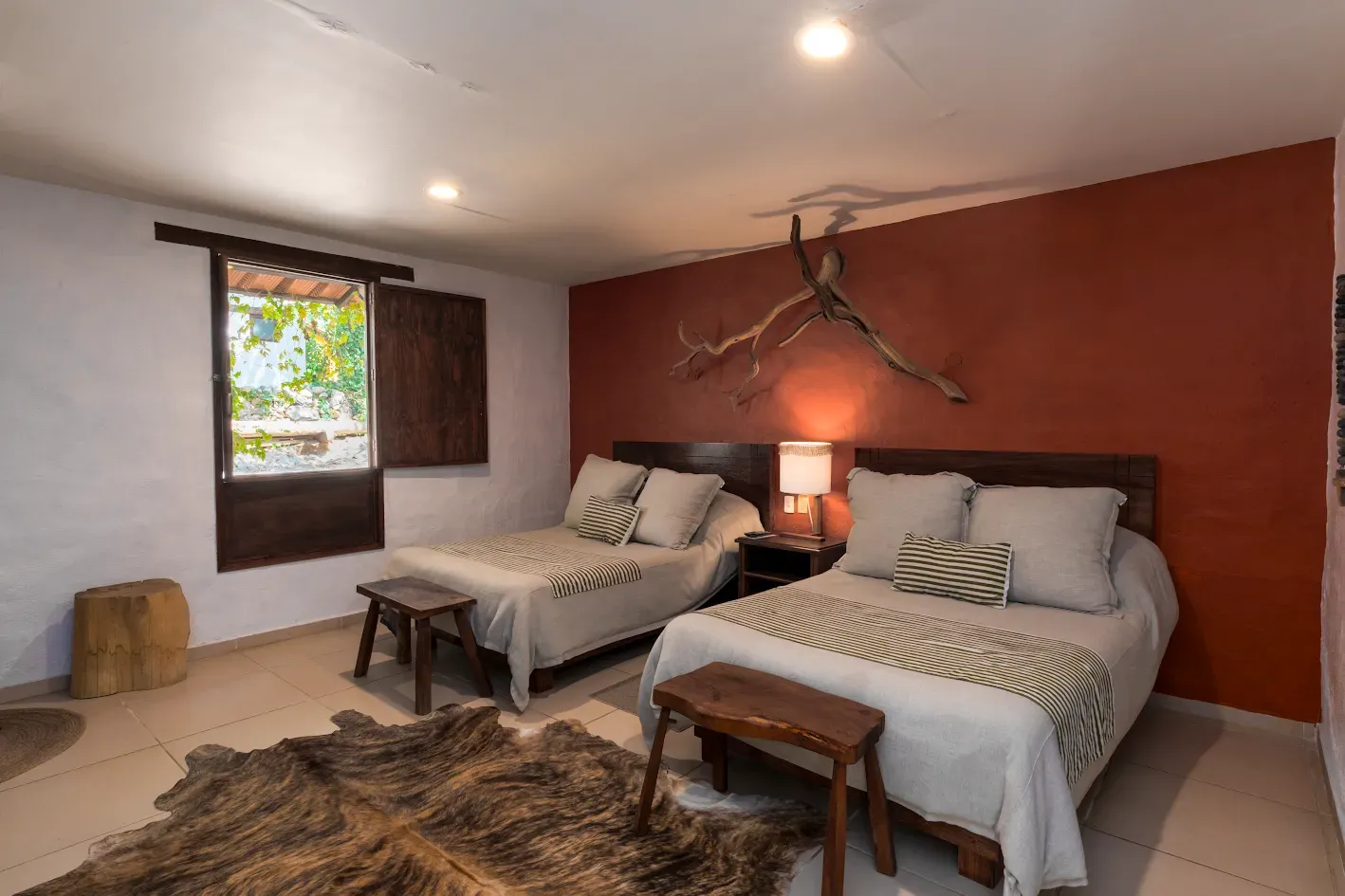 Dormitorio moderno con dos camas individuales, pared decorativa roja, ventana, banco y decoración rústica.