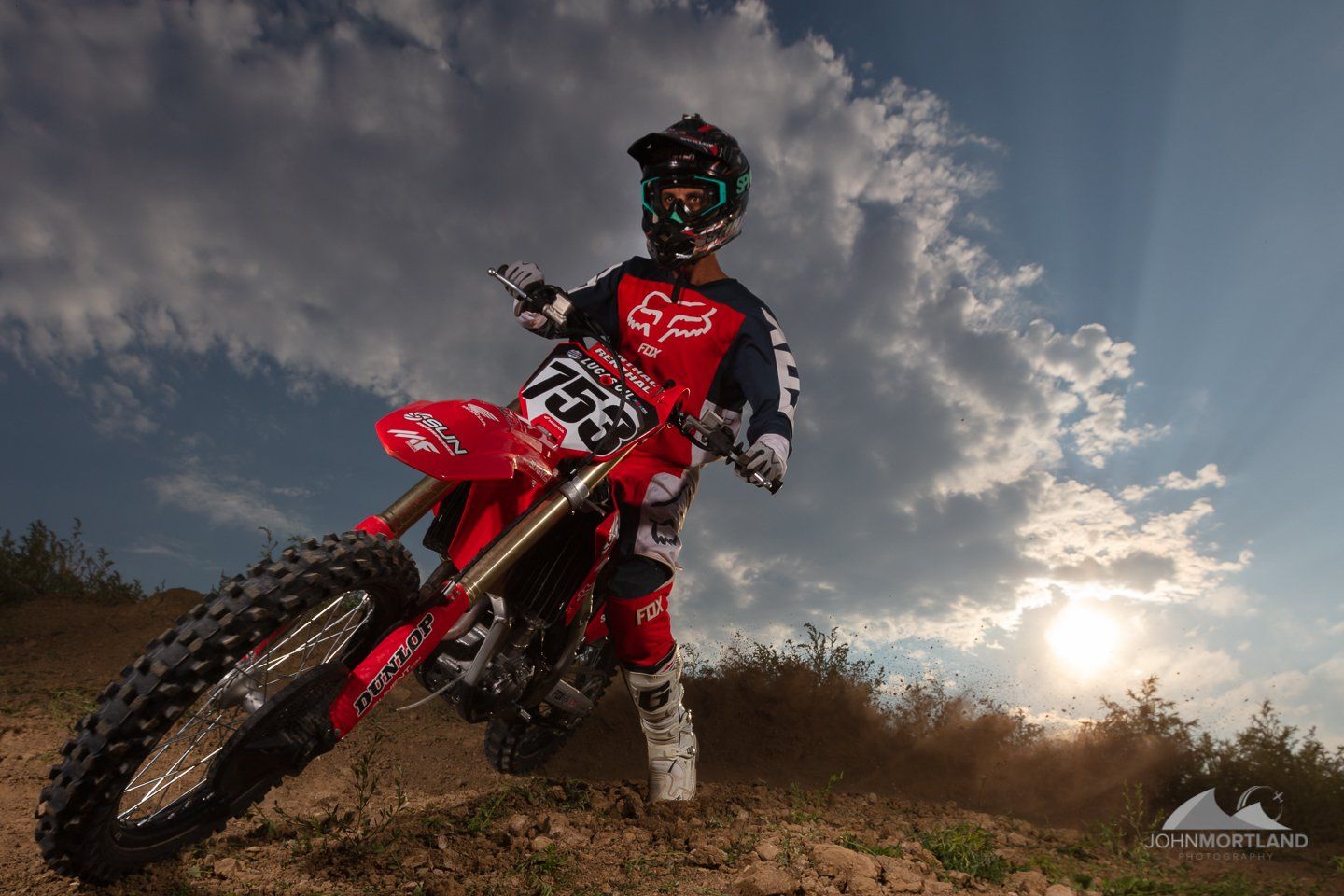Motocross rider on a red motorcycle, poised on a dirt hill. Wearing red and black Fox gear, under a cloudy sky.