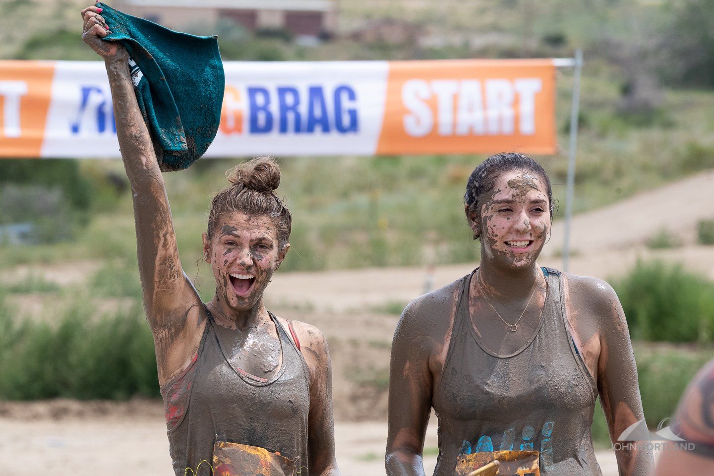 Two mud-covered women celebrate at a race start line, one holding a towel aloft and smiling.