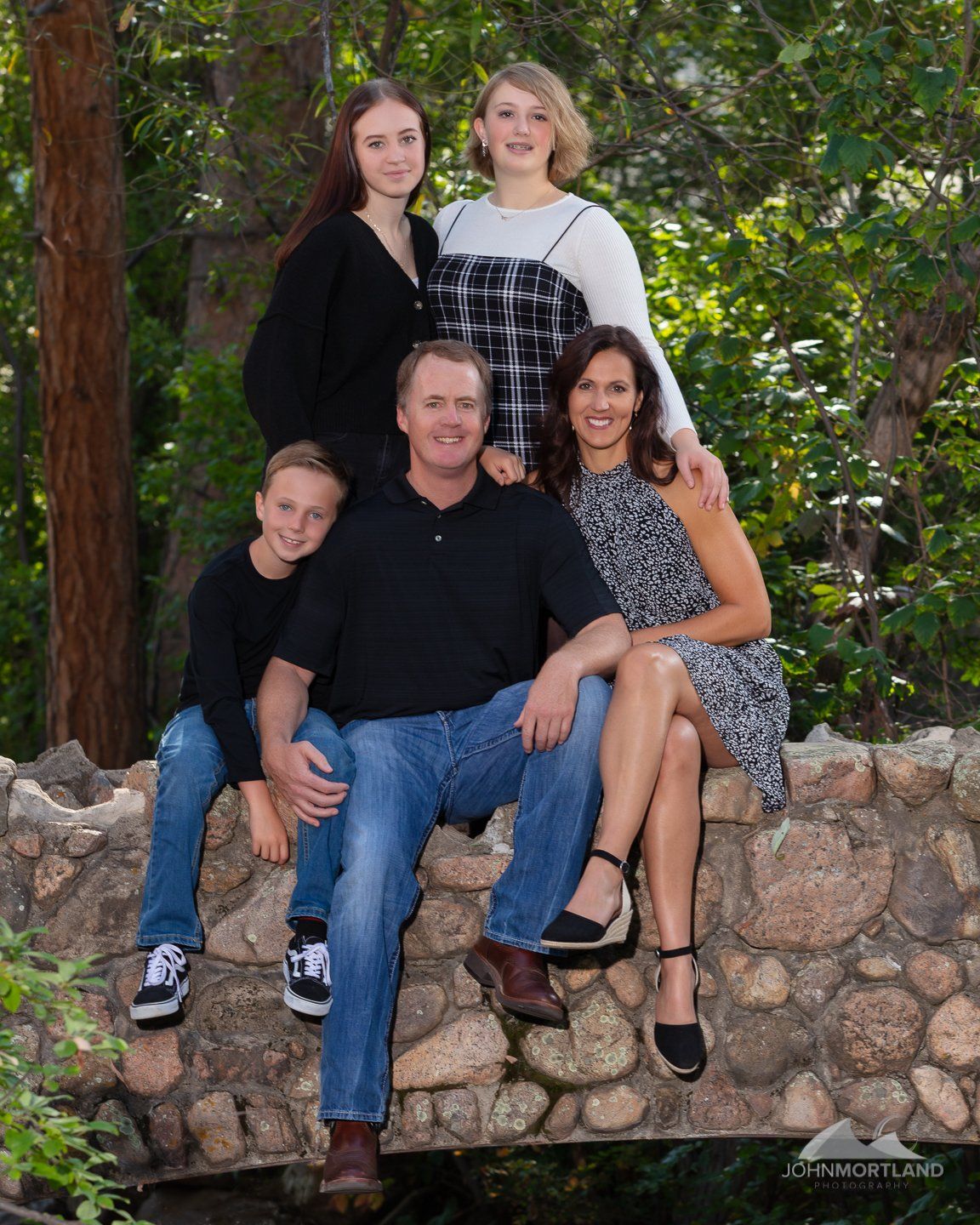 Family of five poses on a stone bridge, smiling. Forest background, natural light.