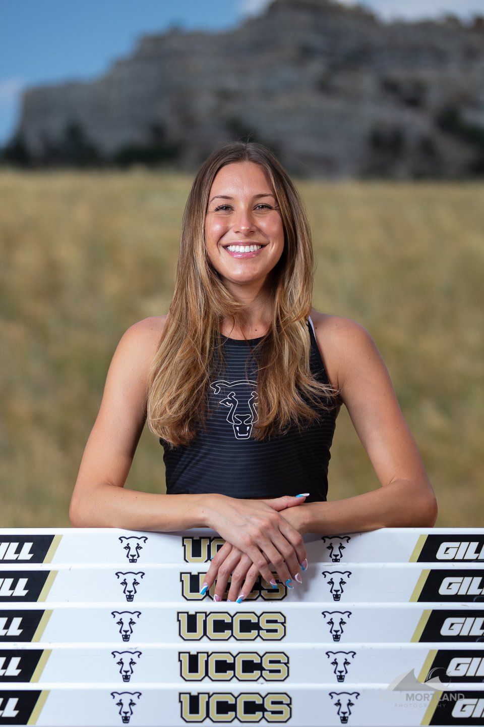 Woman in athletic wear smiles, leans on hurdles with UCCS logo in outdoor setting.