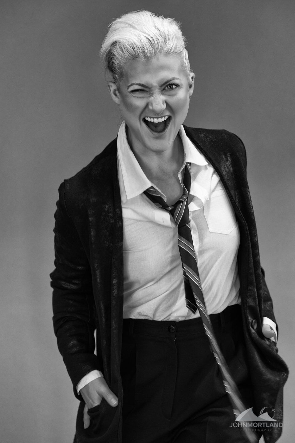 Woman in suit, open-mouthed, laughing. Short, blonde hair, tie askew, hand in pocket. Black and white.