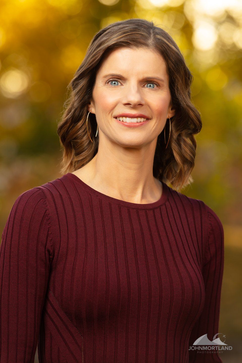 Woman with wavy brown hair smiles at the camera, wearing a maroon sweater with a blurry yellow and green background.