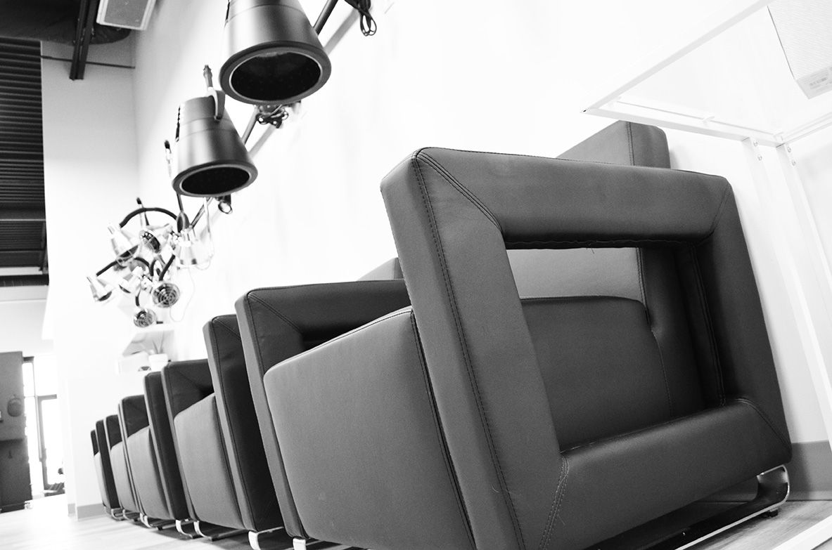 Row of black salon chairs with hooded hair dryers, white walls.