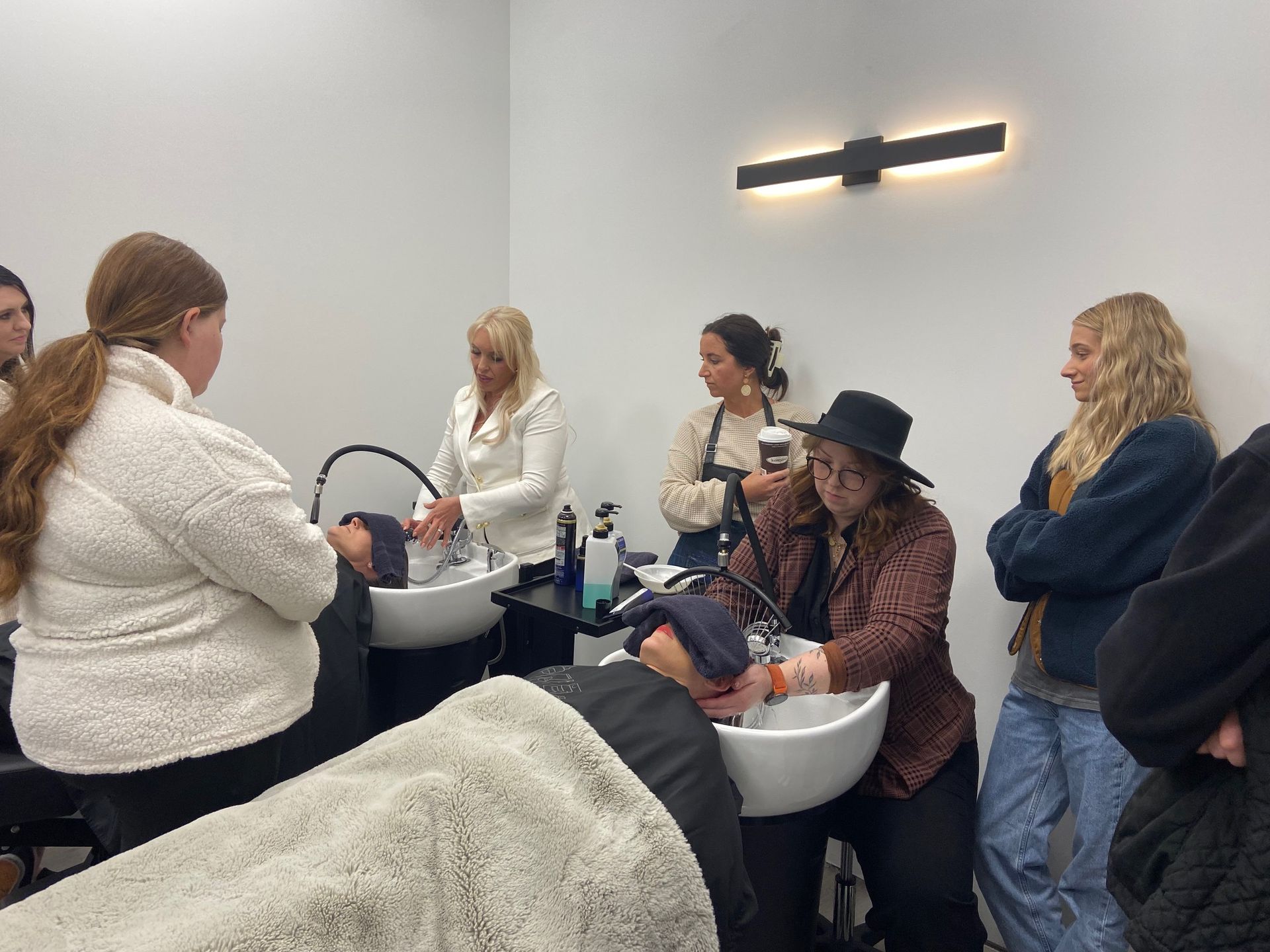 People in a salon setting, one having hair washed at a bowl, others observing, some holding products.