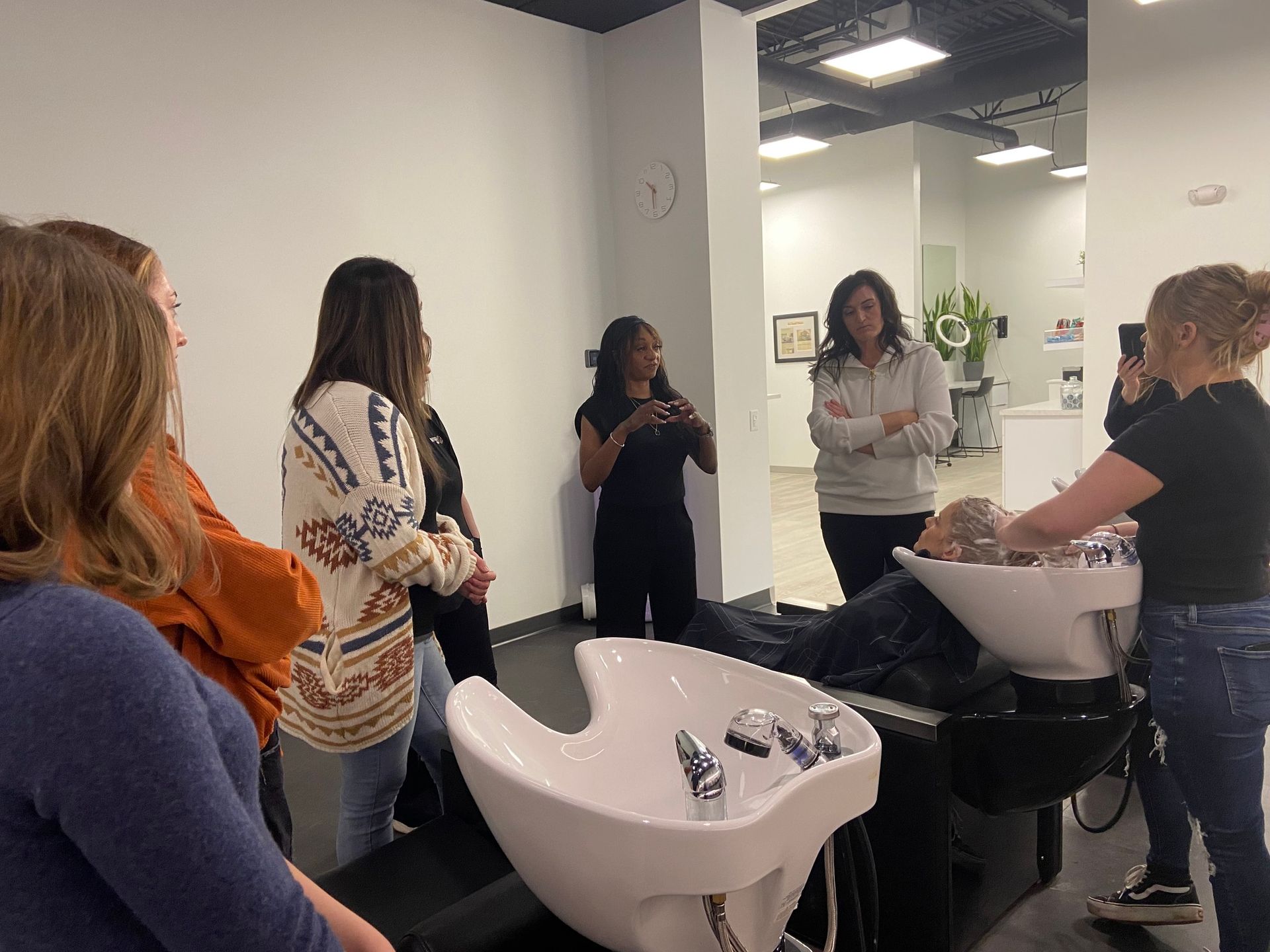 Salon students watching a woman wash a client's hair in a sink.