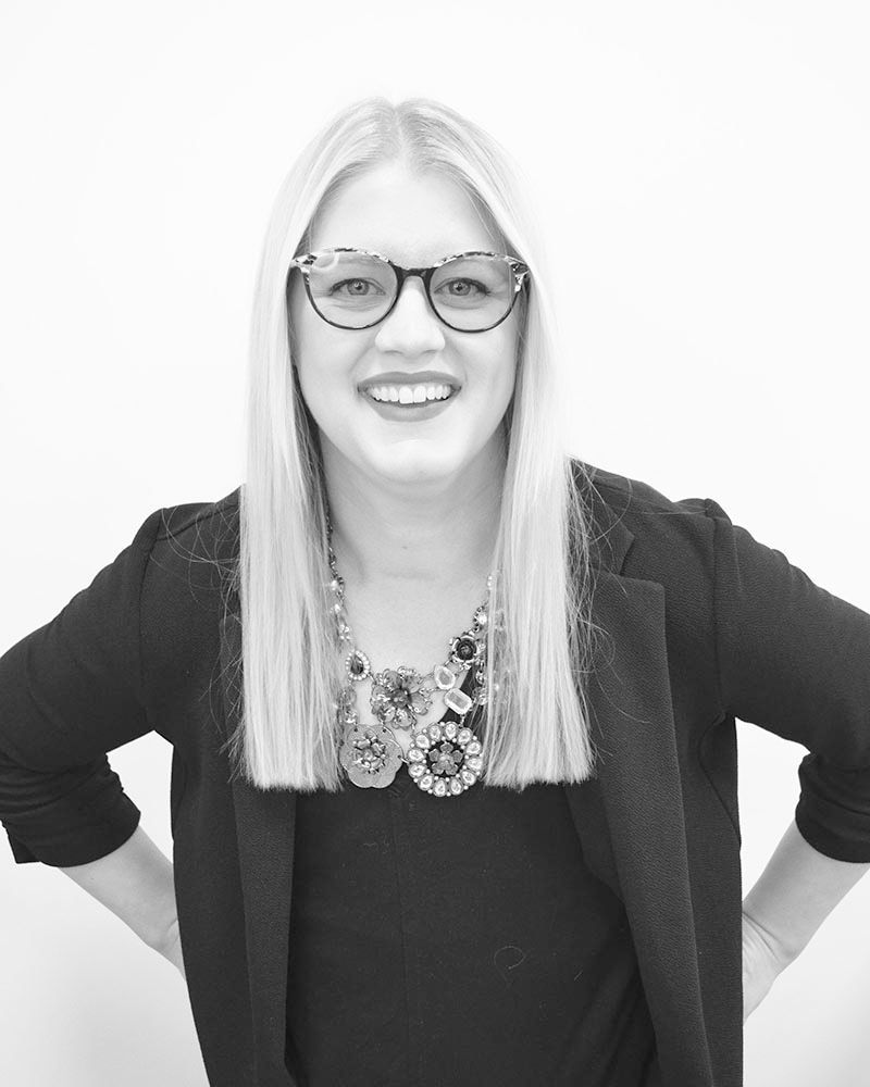 Blonde woman smiling, wearing glasses, blazer, and statement necklace; hands on hips.