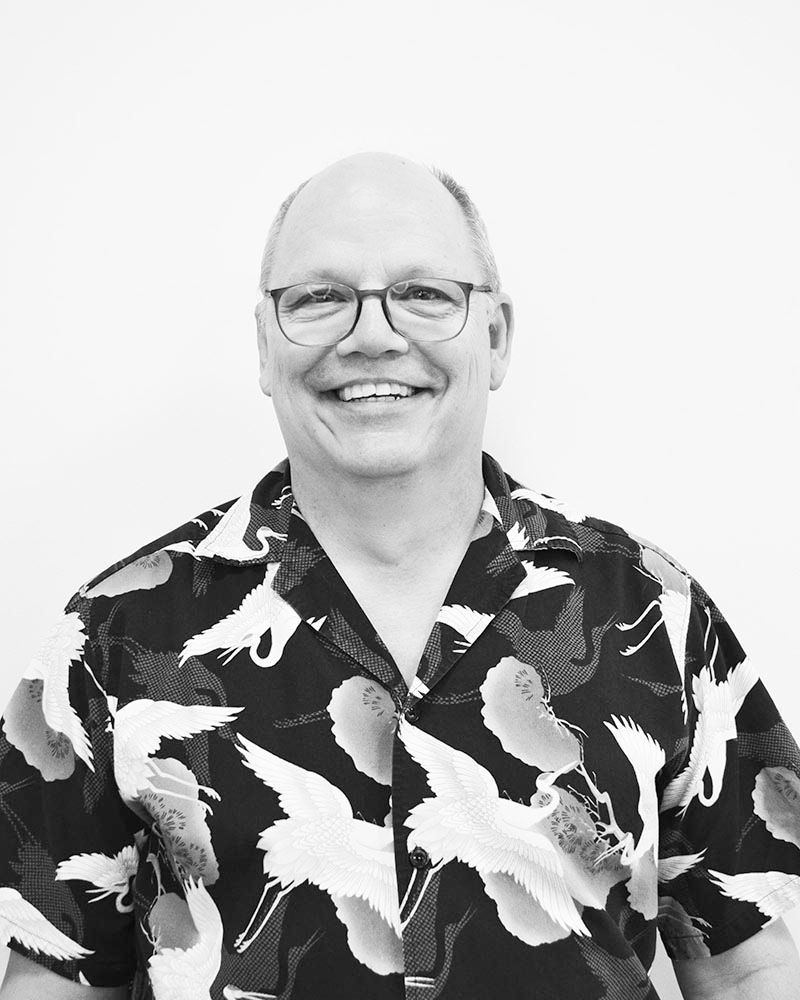 Man with glasses smiling, wearing a black shirt with white bird design; against a white backdrop.