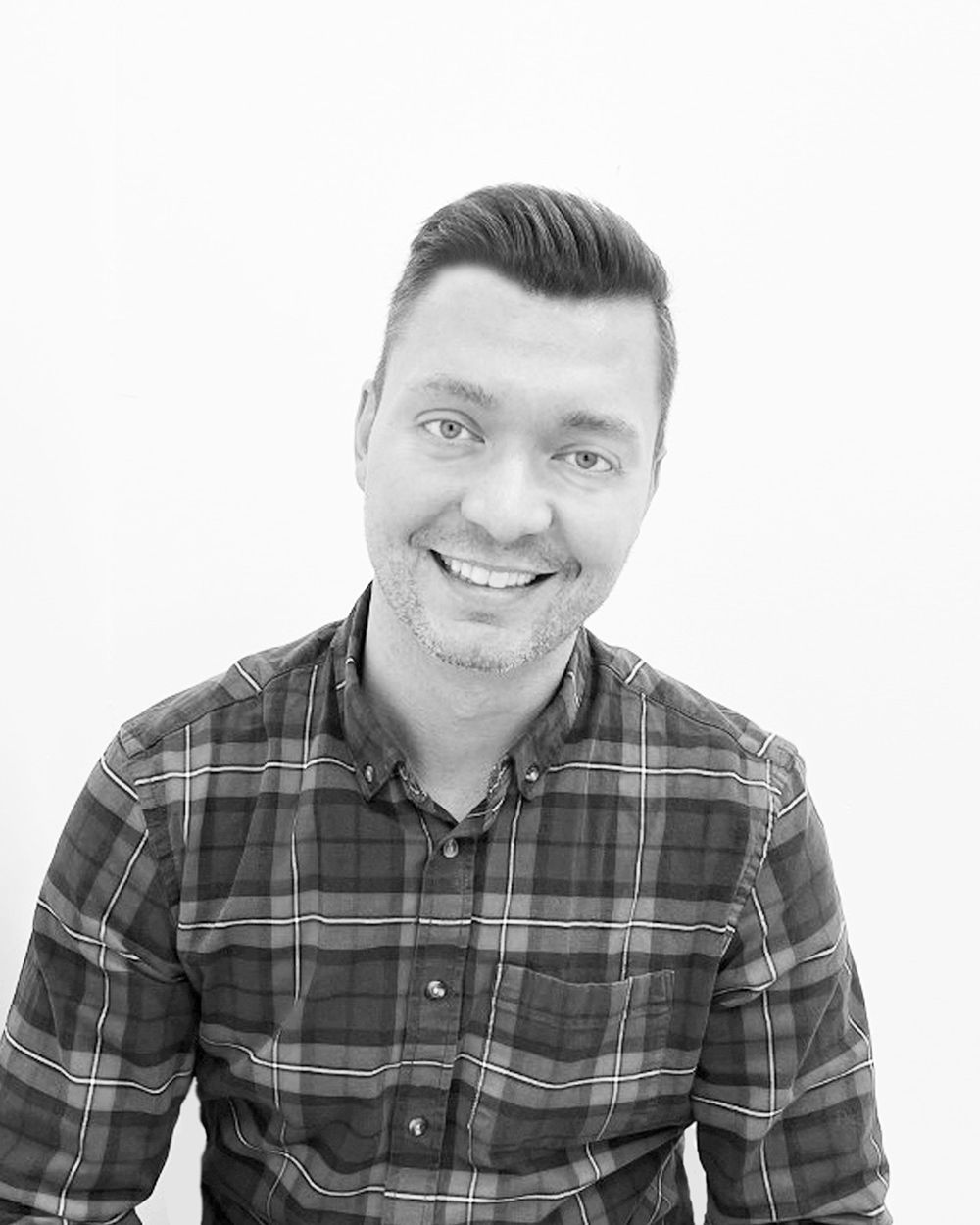 Man with dark hair and a plaid shirt smiles at the camera, against a white backdrop.