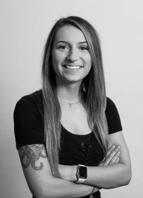 Smiling person with long hair and tattooed arm, standing with arms crossed against a plain background