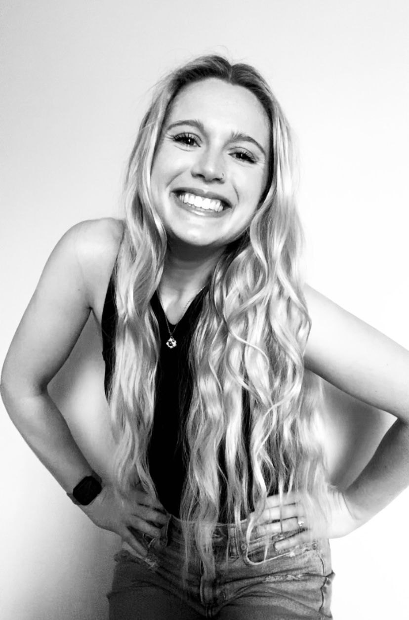 Woman with long wavy hair smiles over her shoulder, wearing a black shirt, against a white wall.