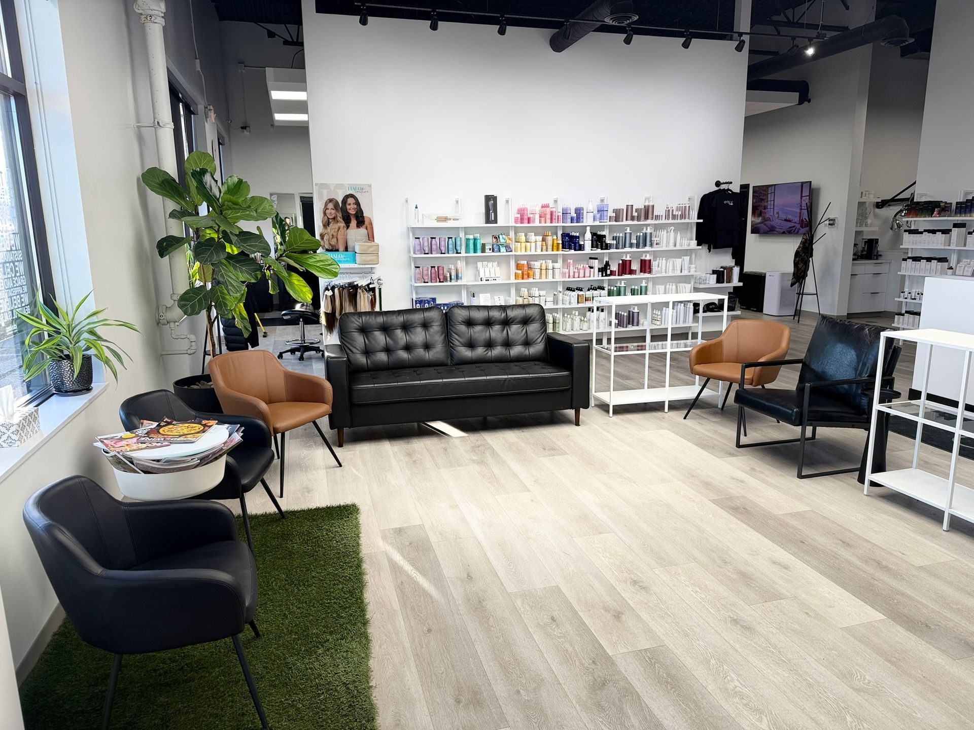 Hair salon waiting area: black sofa, chairs, plant, product display, and a white wall.