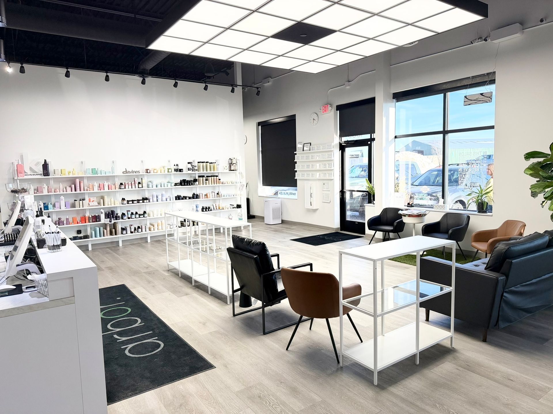 Brightly lit modern retail space with white shelving displaying products; seating area with black and brown chairs; window view of outdoors.