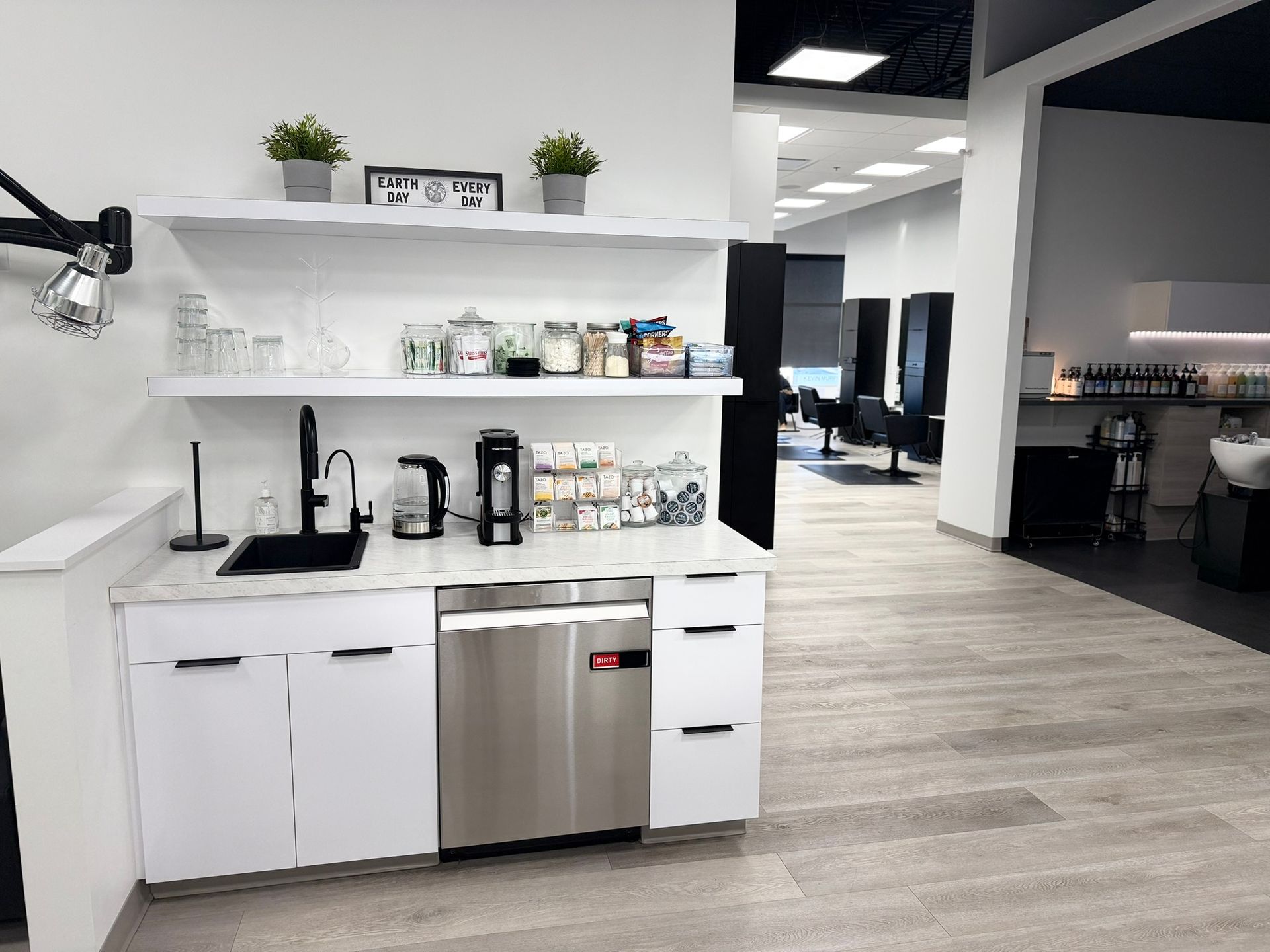 Salon interior with a coffee station. White cabinets, stainless steel appliances, shelves with supplies, and light wood flooring.