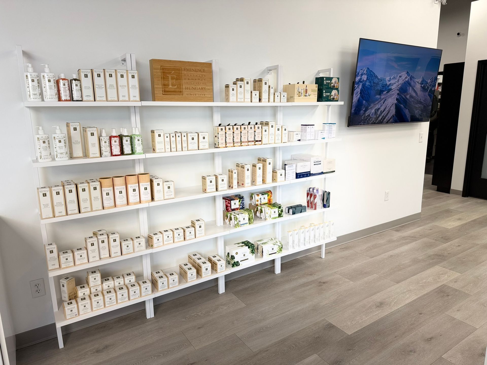 White shelves stocked with various products against a white wall; a TV displays a mountain landscape.