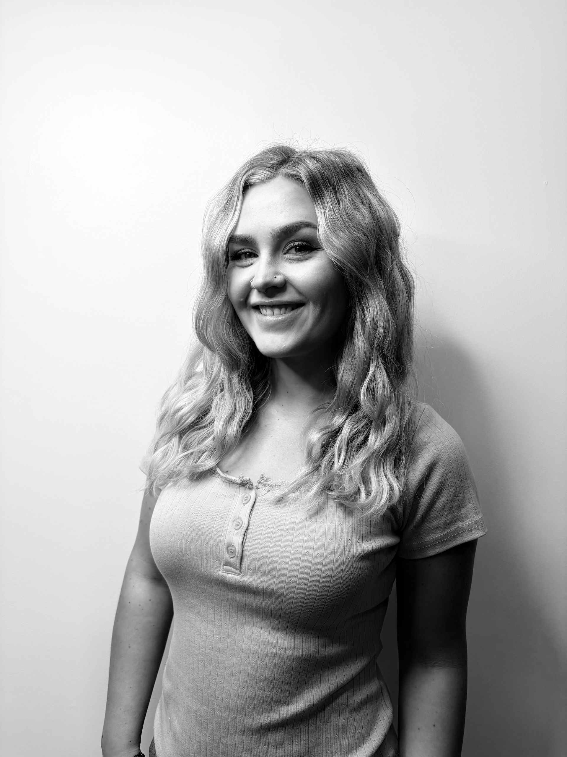 Woman with long blonde hair smiles, wearing a button-up shirt over a white turtleneck, set against a plain background.