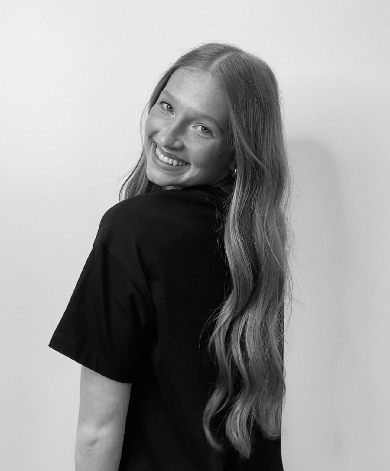 Woman with long wavy hair smiles over her shoulder, wearing a black shirt, against a white wall.