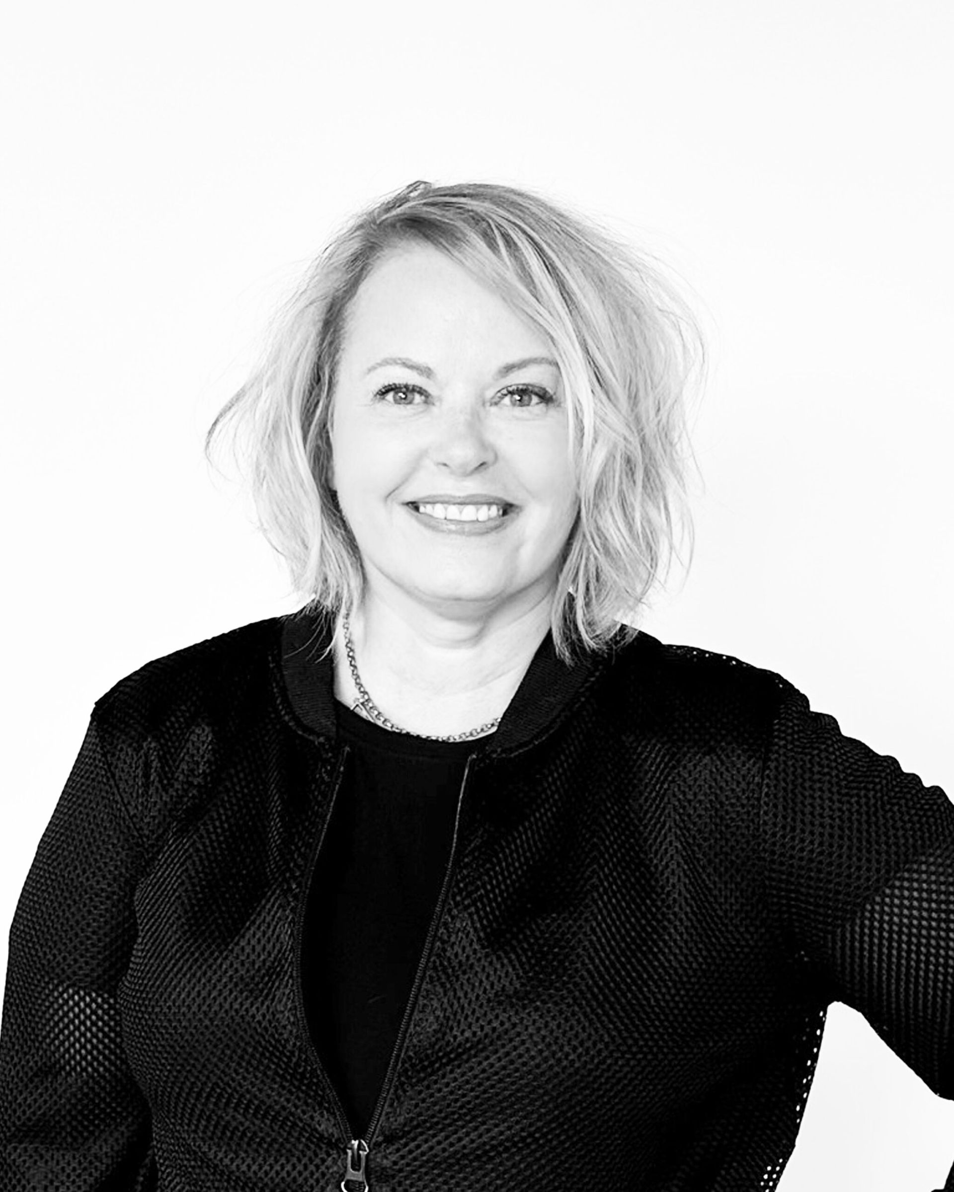 Woman smiling, wearing a black jacket and necklace, in front of a white background.