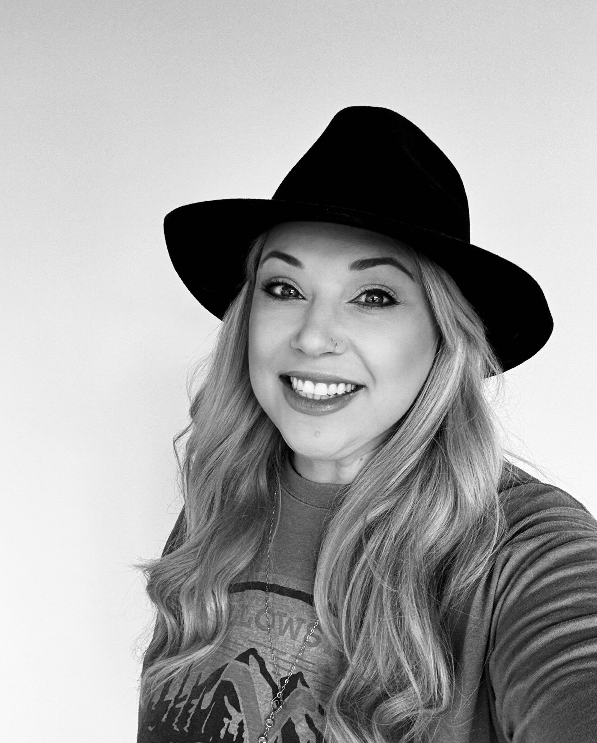 Woman with long blonde hair, wearing a black hat and smiling, in front of a white background.