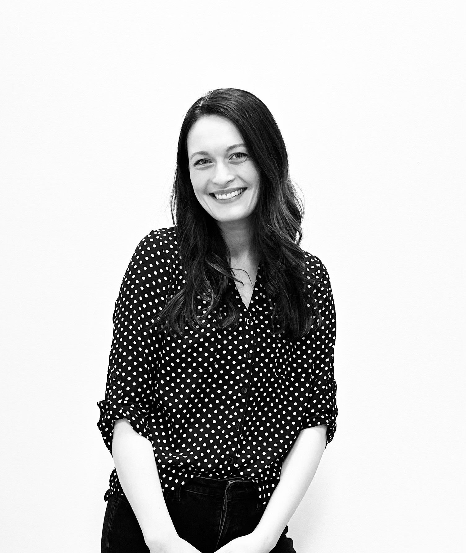 Woman with long dark hair, smiling, wearing a polka-dot blouse, against a white background.