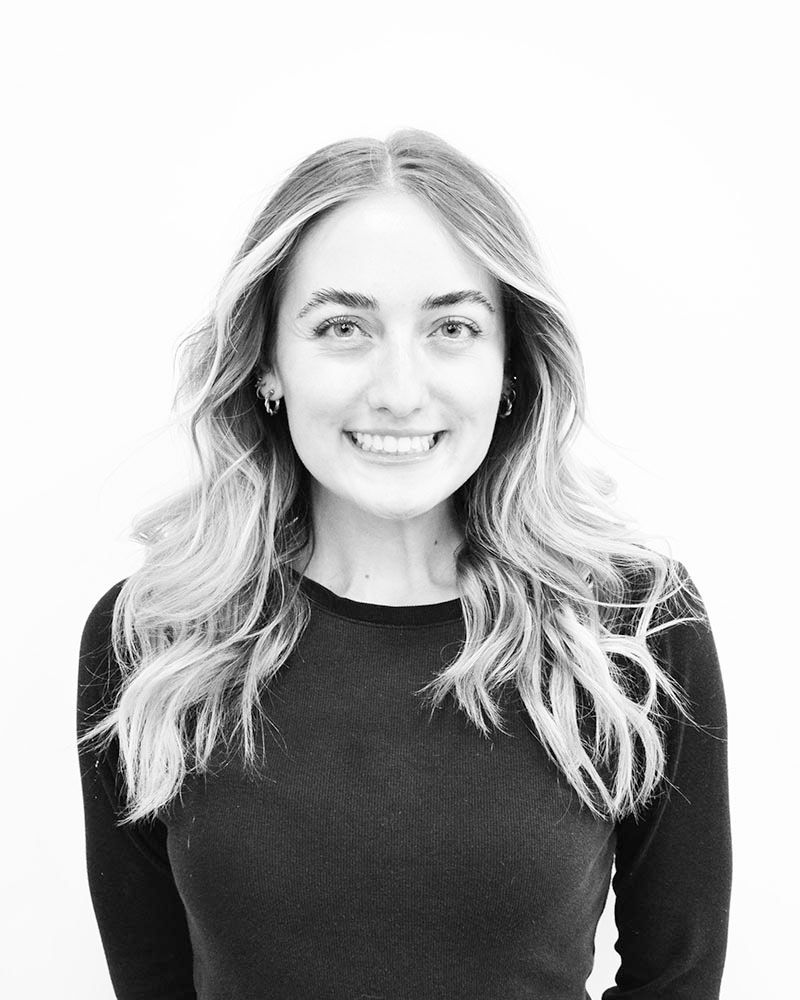 Woman with long, wavy blonde hair smiles, wearing a black sweater, against a white backdrop.