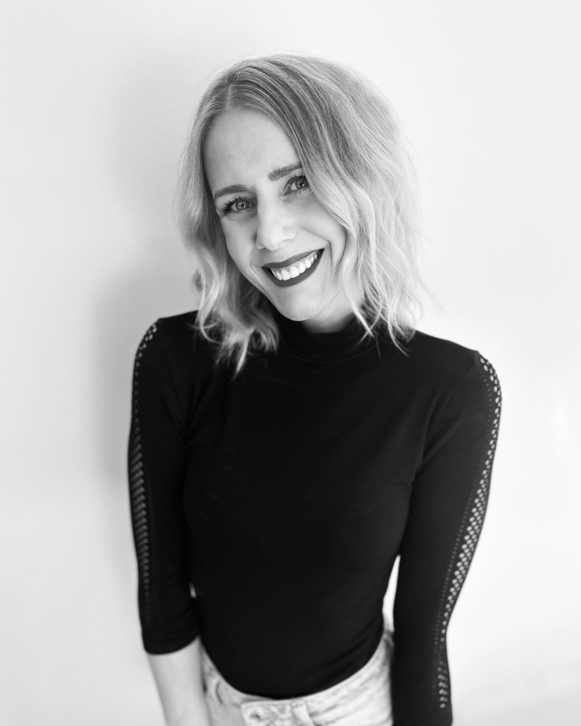 Woman with blonde wavy hair, smiling, wearing a black turtleneck and light-wash jeans.