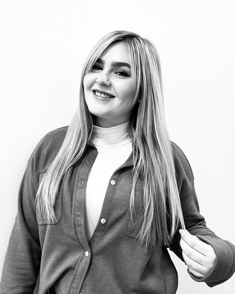 Woman with long blonde hair smiles, wearing a button-up shirt over a white turtleneck, set against a plain background.