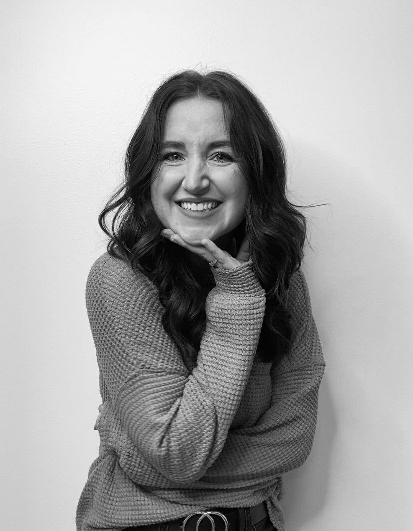 Woman smiling, hand near face, wearing a sweater, standing in front of a white wall.