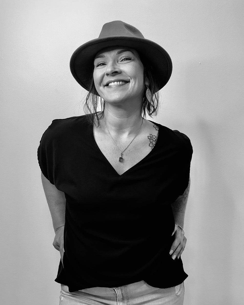 Woman in a fedora and black top smiling, hands in pockets.