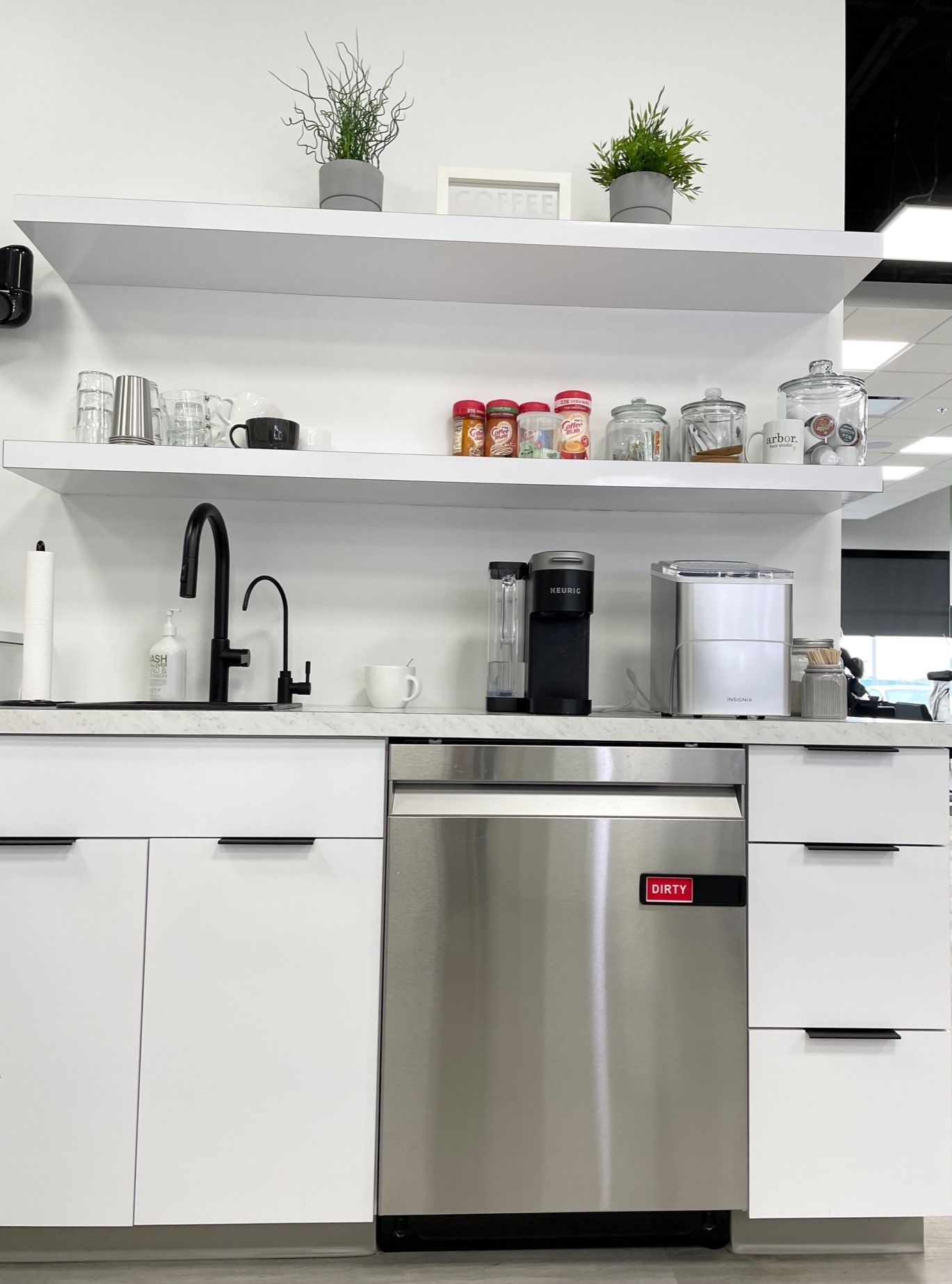 White office kitchen with a sink, stainless steel dishwasher, and shelves with plants and supplies.
