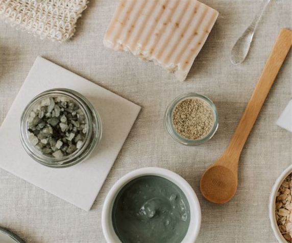 A collection of natural skincare ingredients: a clay mask, bath salts, a bar of soap, and dry oats on a linen surface.