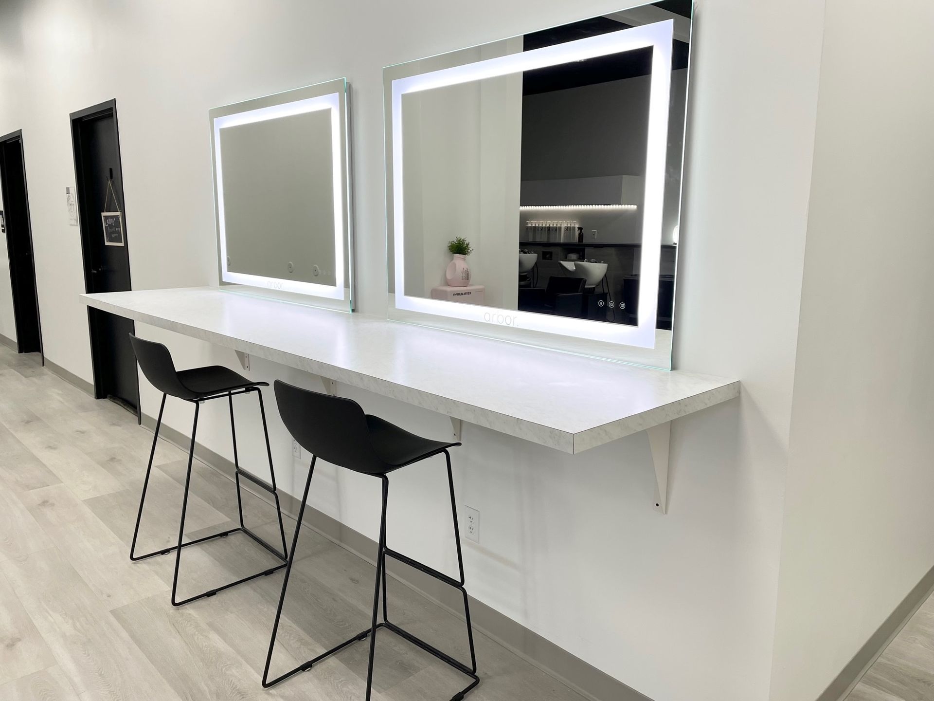 Two illuminated mirrors above a white counter with black barstools in a modern salon.