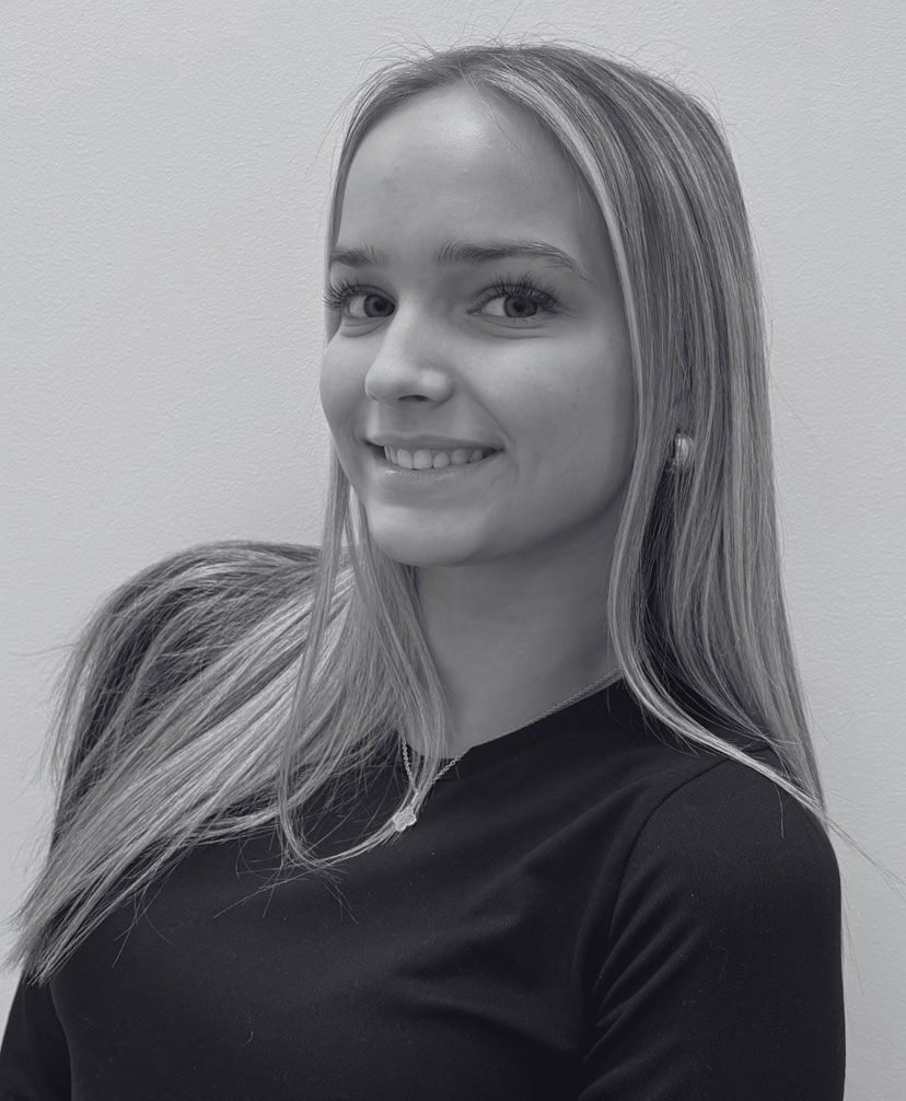 Woman with long blonde hair, smiling, wearing a black shirt, leaning against a white wall.
