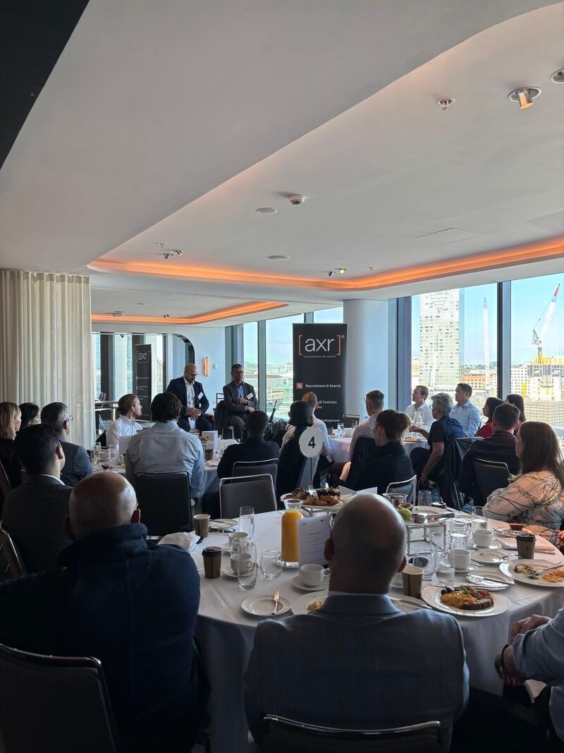 A business presentation in a bright, modern room with floor-to-ceiling windows, showing attendees seated at round tables.