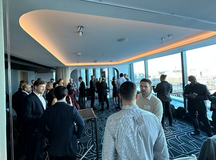 A group of people in business casual attire networking in a modern, high-rise room with floor-to-ceiling windows.