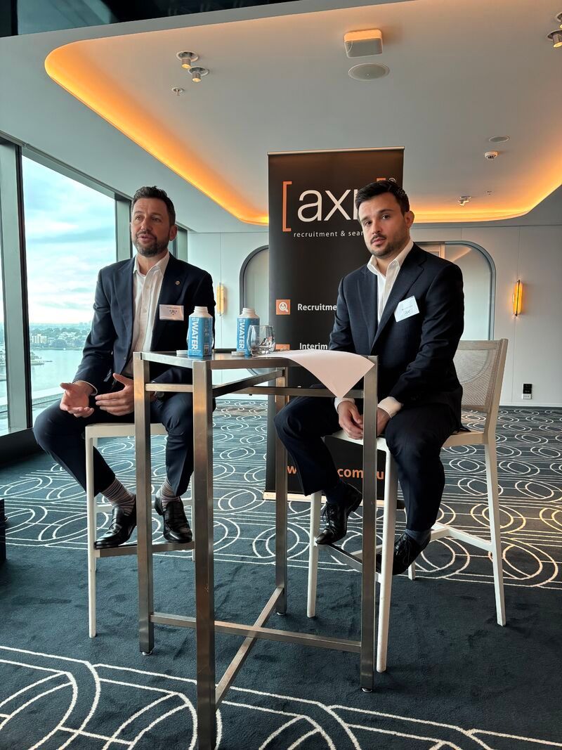 Two people in business suits sit at a high table during a professional event in a modern room with a city view.
