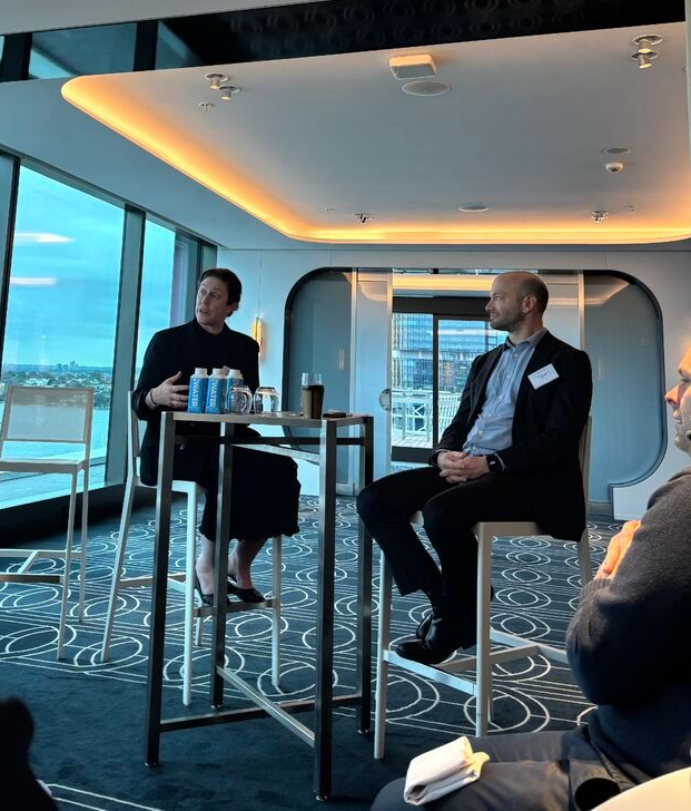 Two people sit at a high table in a modern lounge with large windows, engaged in a professional conversation.