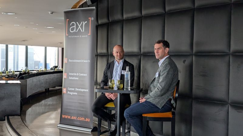 Two professionals sit at a small table for an AXR event, positioned in front of a branded banner in a modern office.