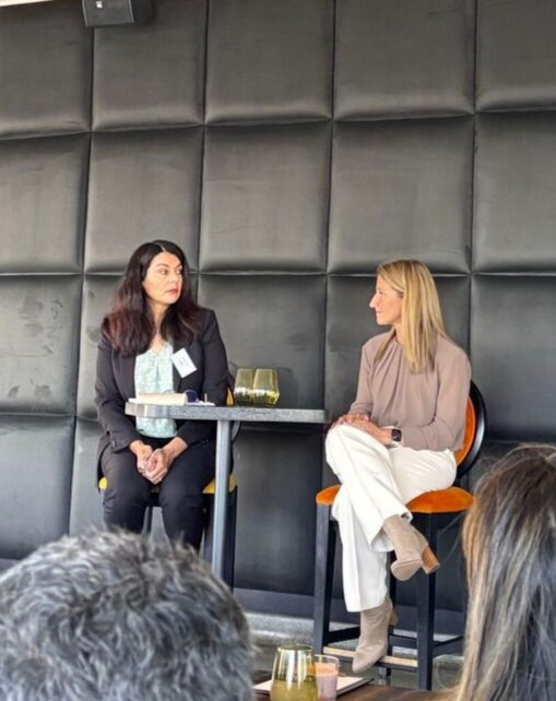 Two people in professional attire sit on chairs during a discussion in front of a dark, paneled wall.