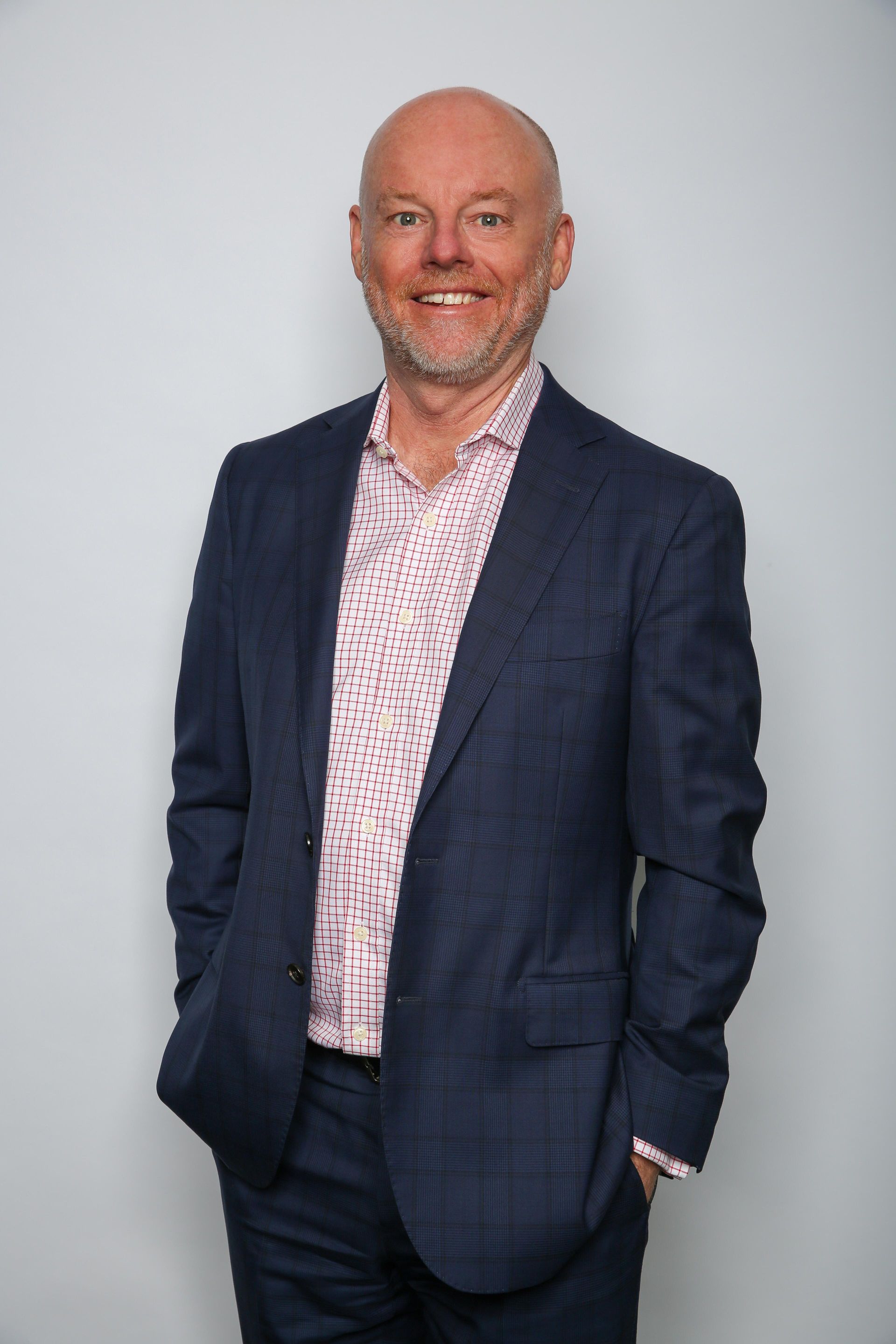 A smiling person in a blue suit and a patterned light-colored shirt stands against a plain grey background.