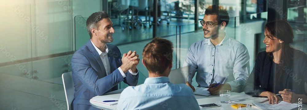 Four colleagues sit around a conference table in a modern glass-walled office, engaged in a professional discussion.