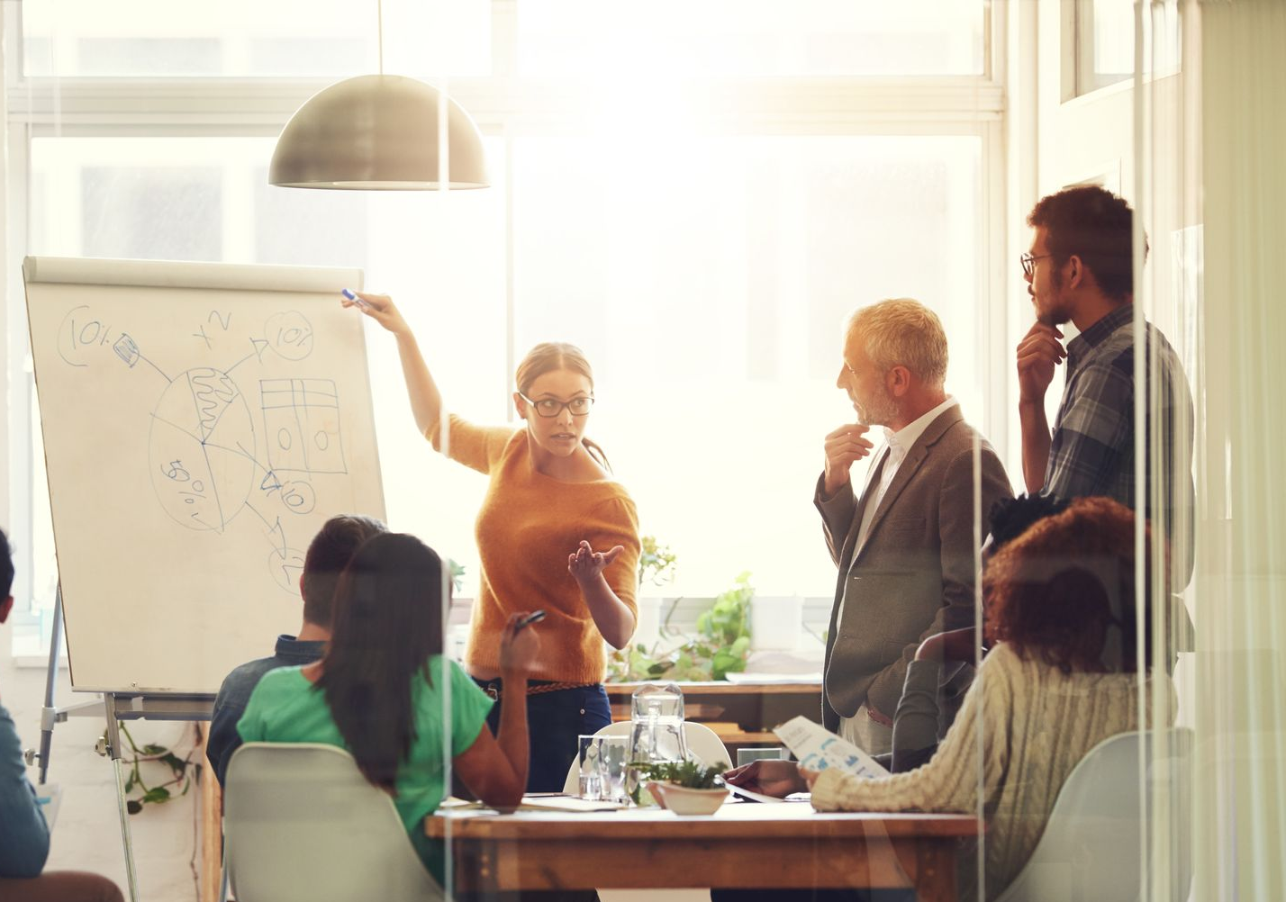 Team meeting in a bright office, with one person presenting at a whiteboard and others listening.