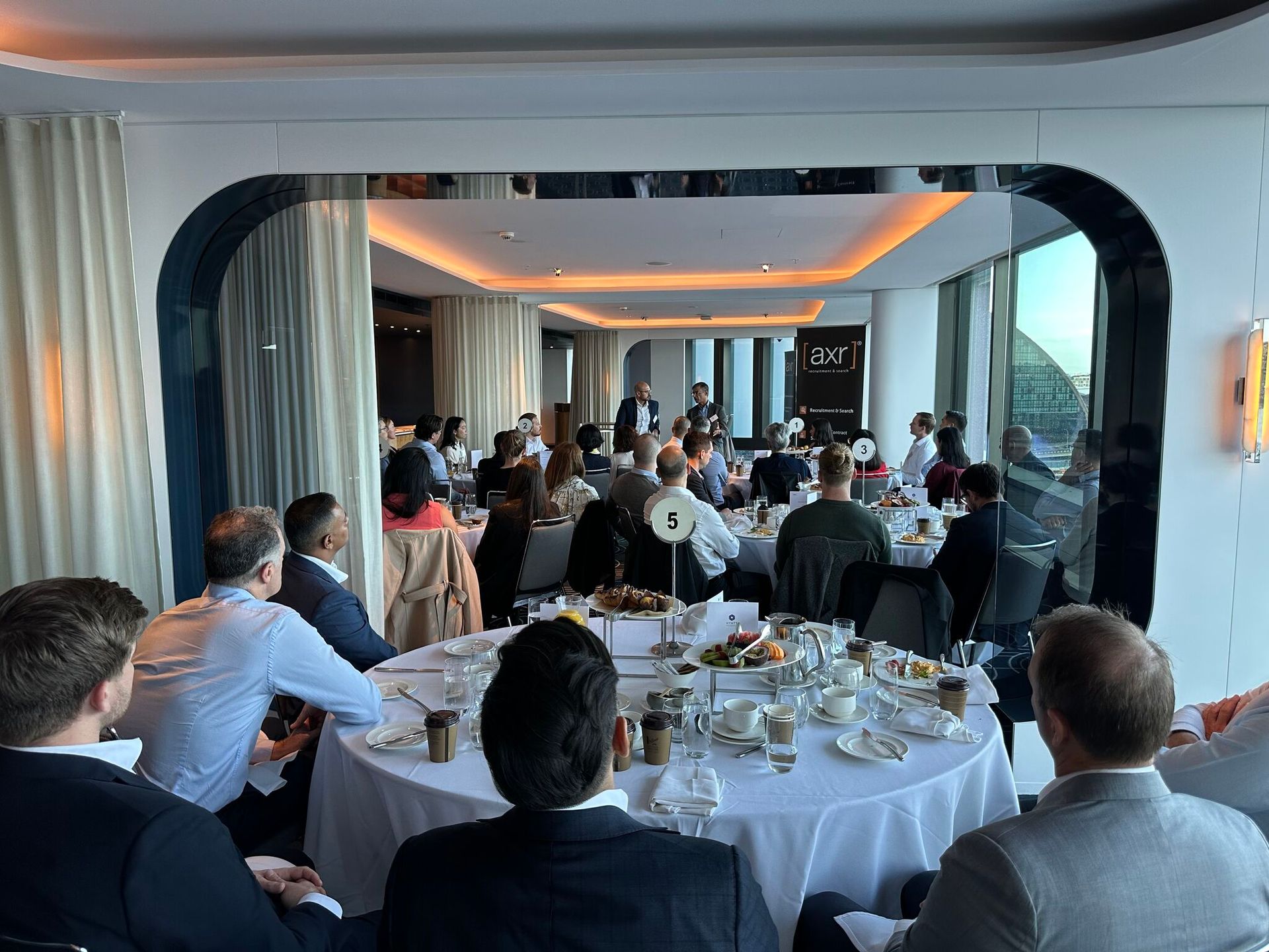 A large group of people in business attire sit at round tables in a brightly lit, modern event space during a presentation.