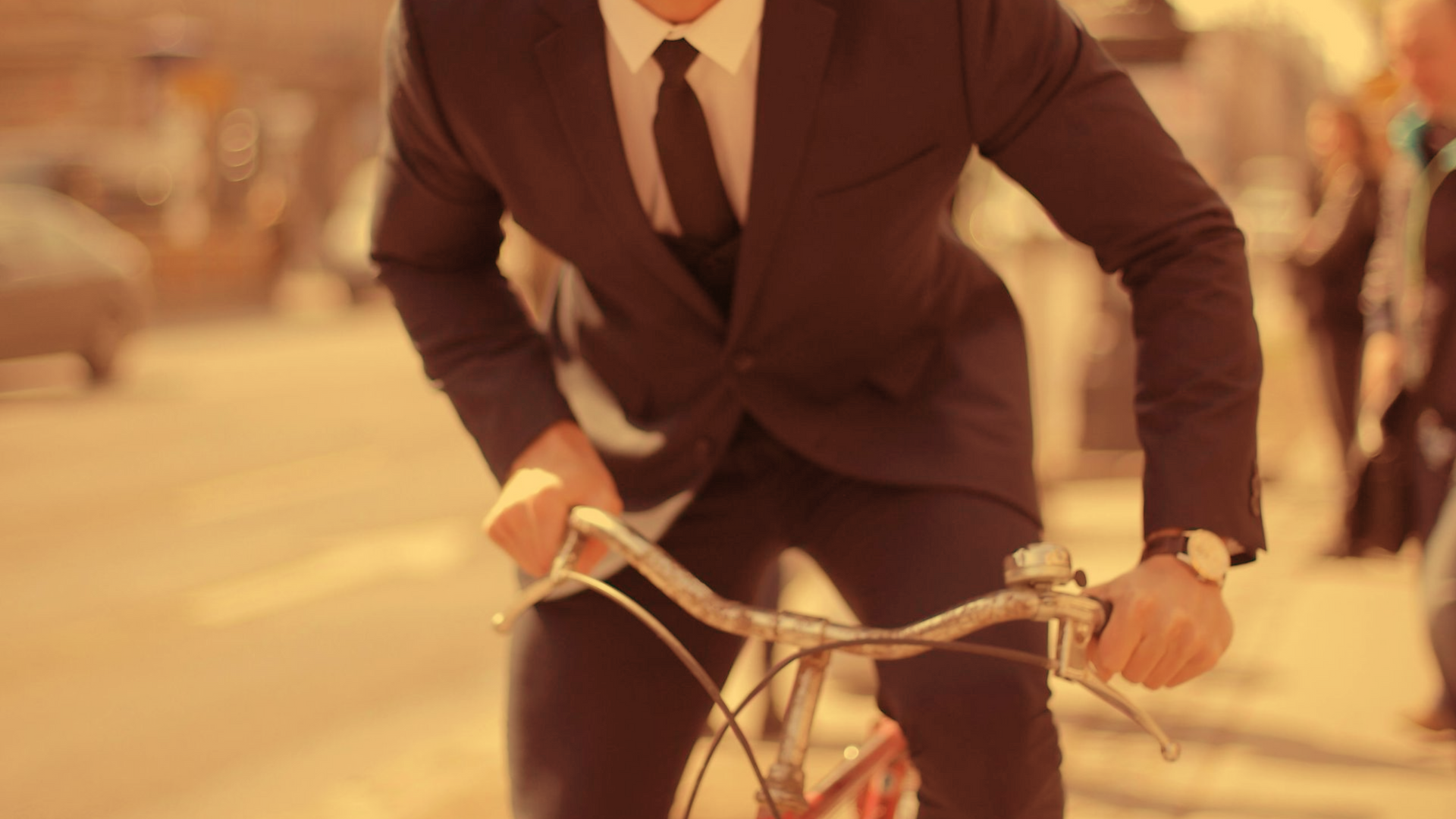 A person in a suit and tie riding a bicycle on a sunlit street, leaning forward while gripping the handlebars.
