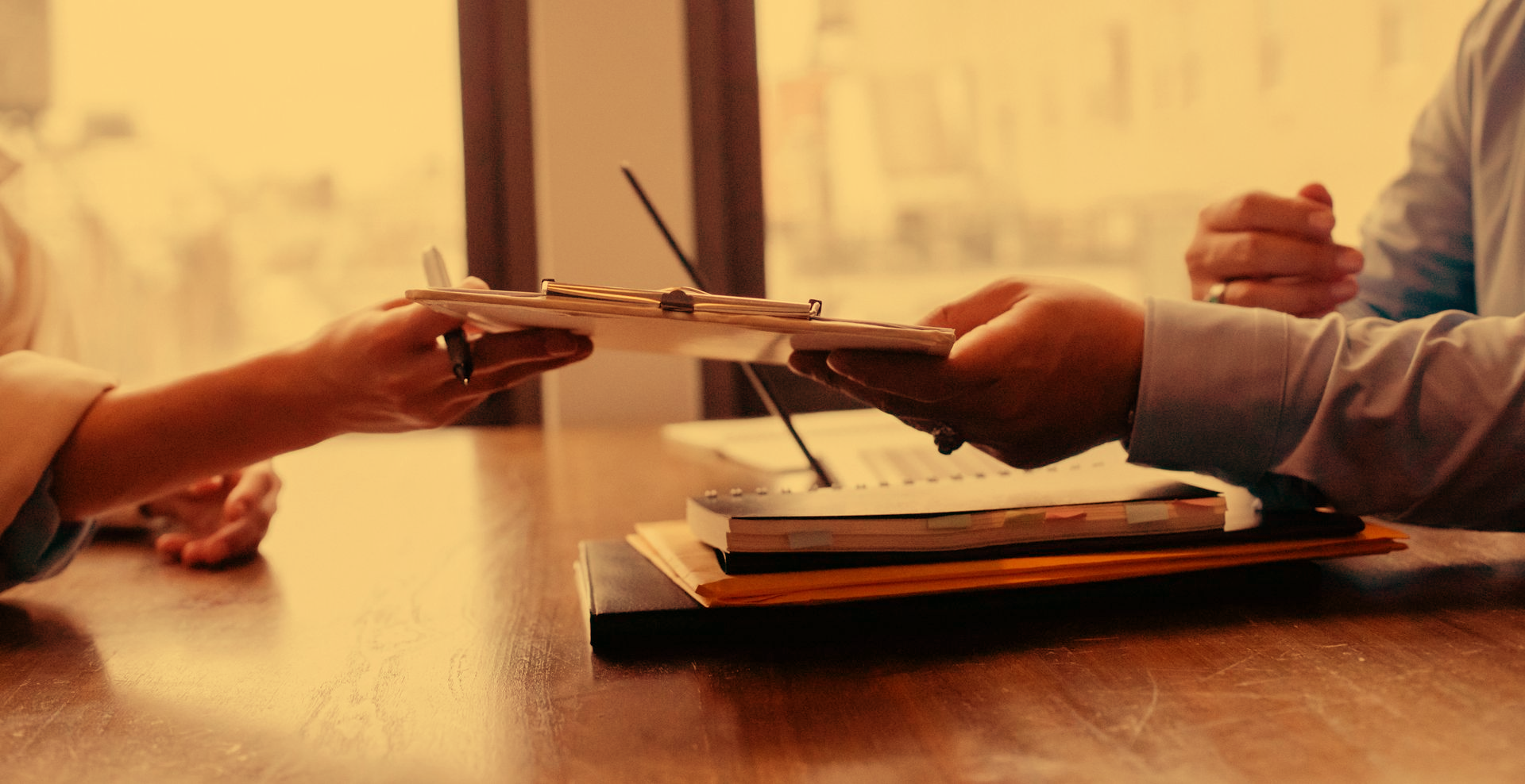Two people sit across from each other at a desk, one handing a document clipboard to the other.
