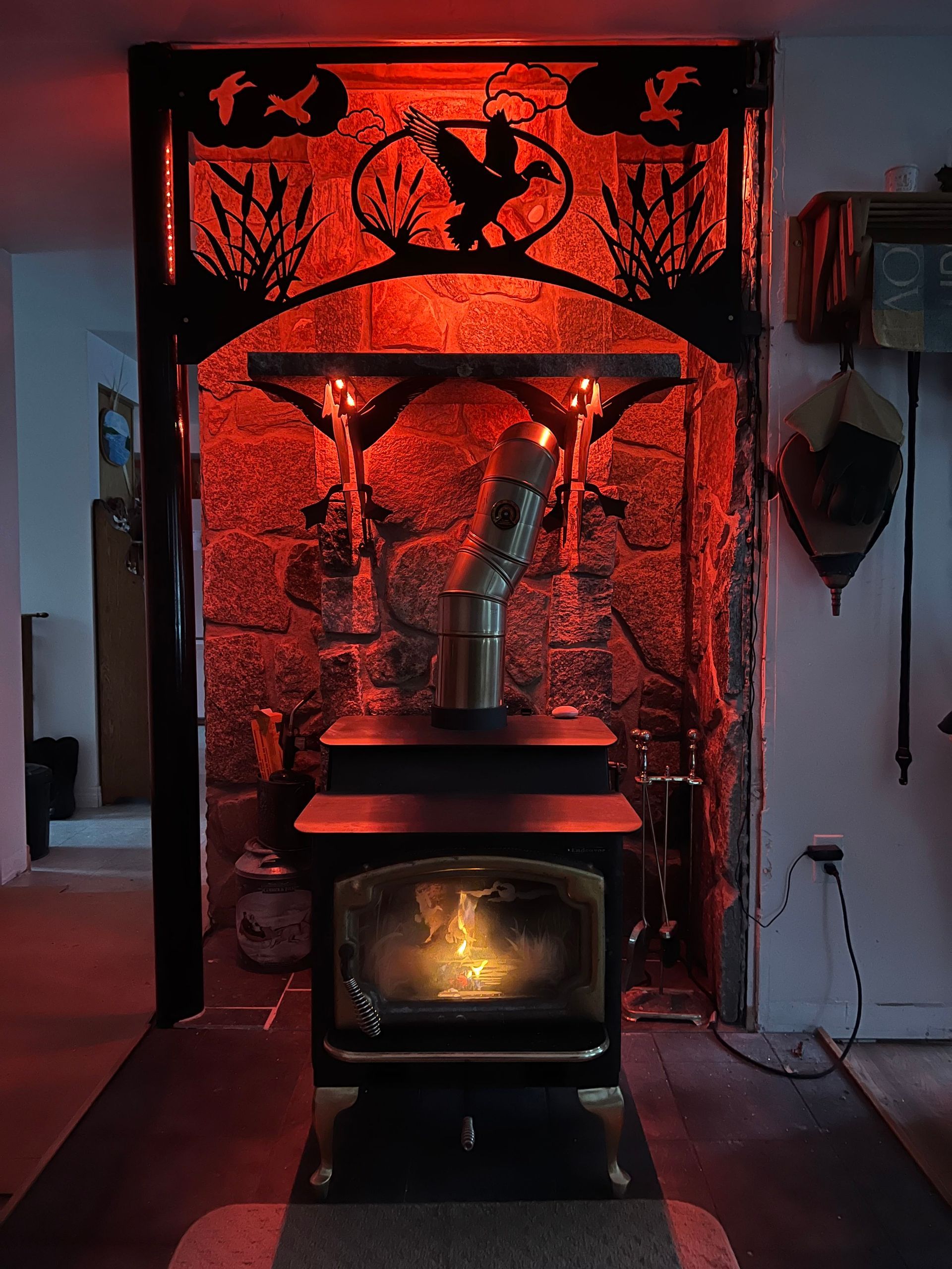 A wood-burning stove sits in a stone hearth framed by a decorative metal screen featuring ducks, glowing in red light.