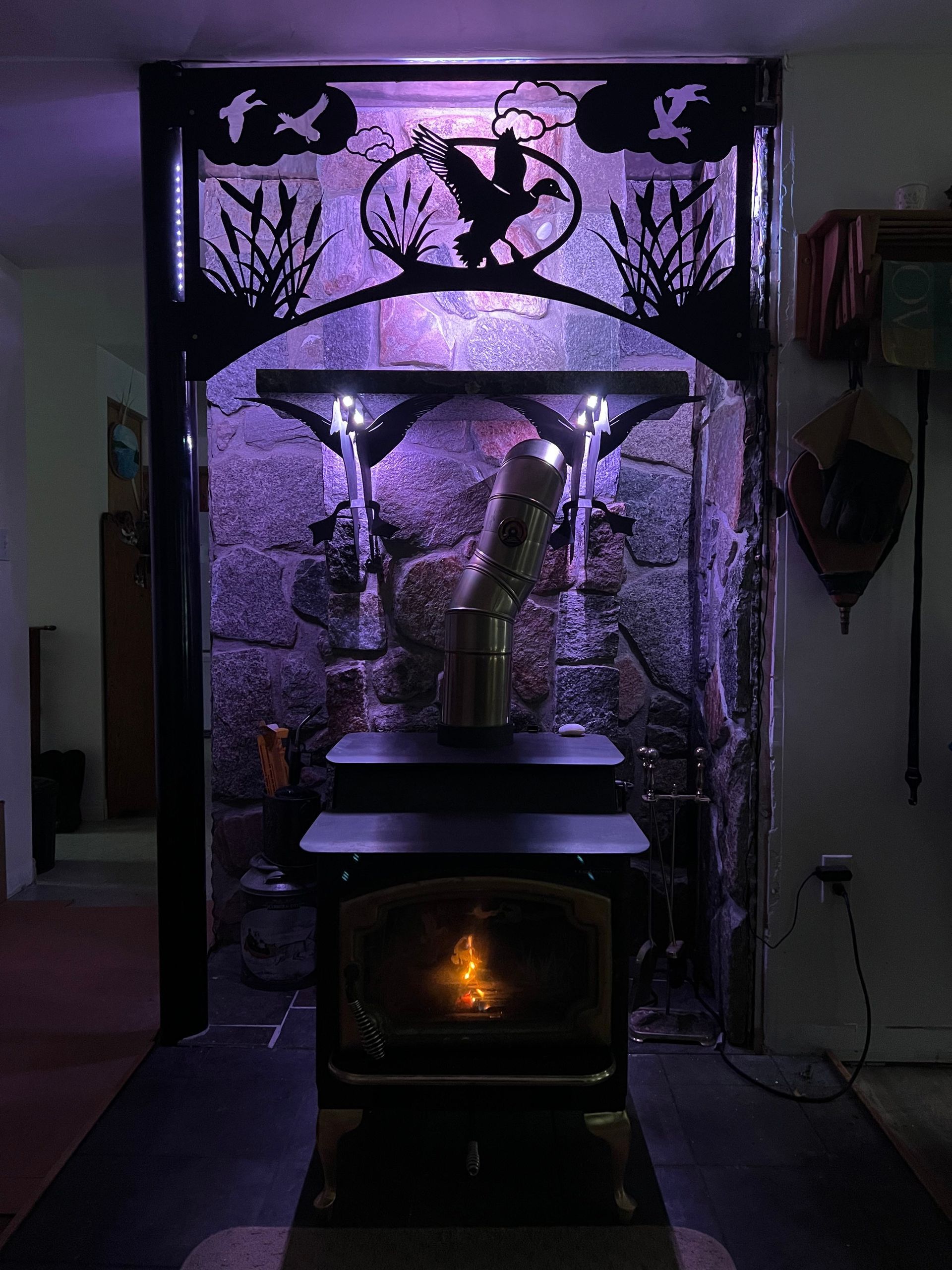 A wood stove sits in a stone-walled hearth illuminated by purple light, topped by a metal duck-themed decorative arch.