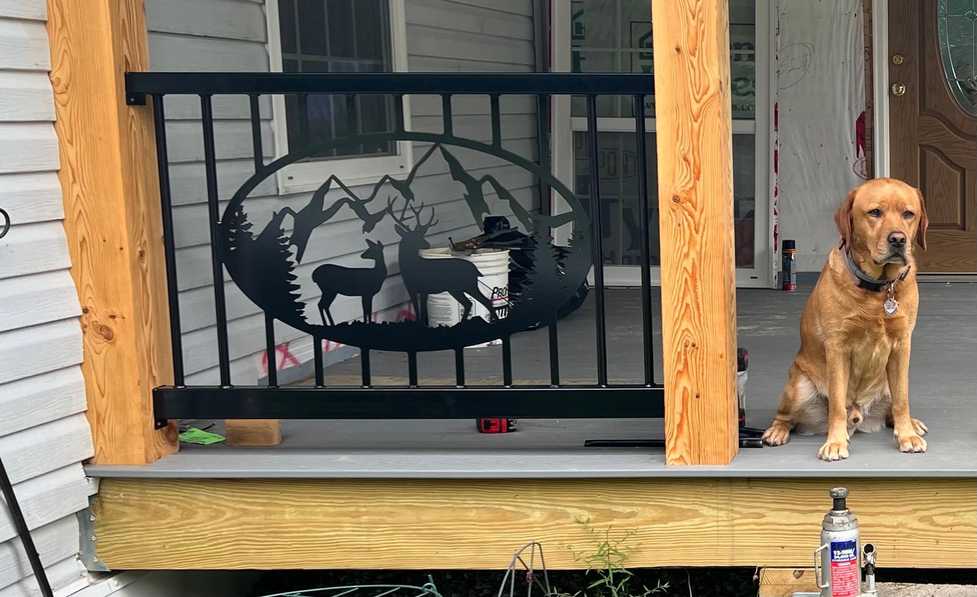 A yellow Labrador sits on a porch next to a black metal railing featuring an oval cutout of mountains and deer.