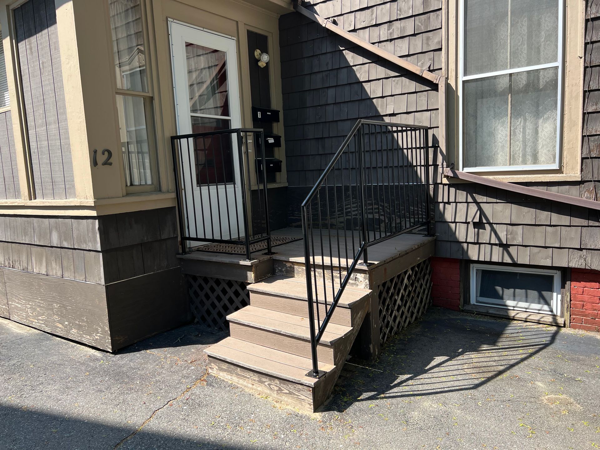 An exterior view of a brown shingled building with a set of wooden stairs leading to a white door with a black railing.